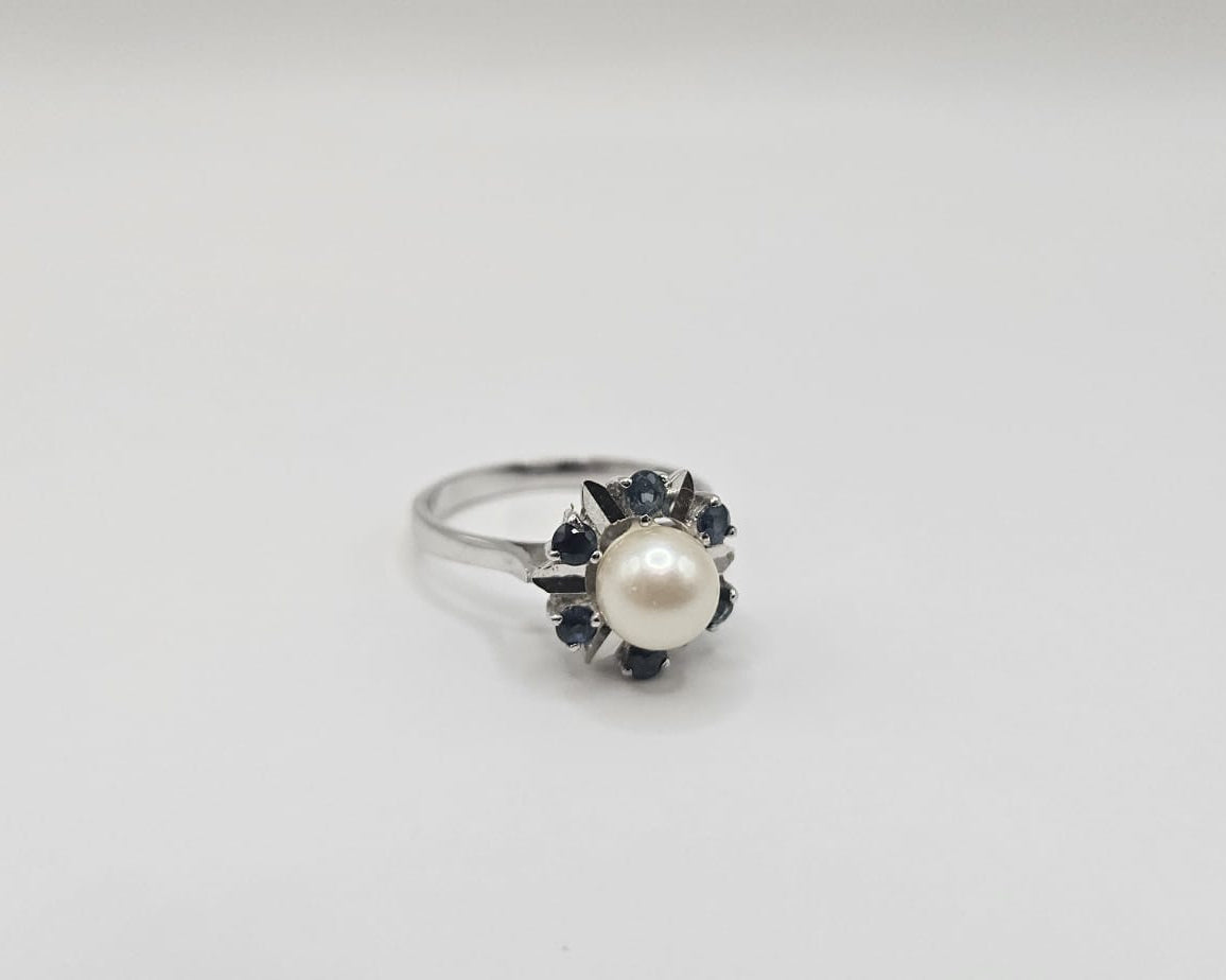 Bague en or blanc ornée d’une perle de culture ronde, entourée de saphirs bleus formant un motif floral élégant, présentée sur fond uni beige clair