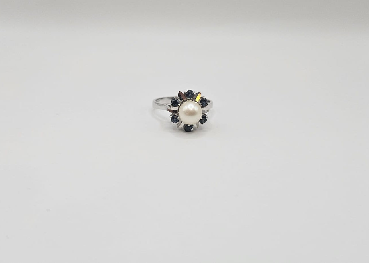 Bague en or blanc ornée d’une perle de culture ronde, entourée de saphirs bleus formant un motif floral élégant, présentée sur fond uni beige clair